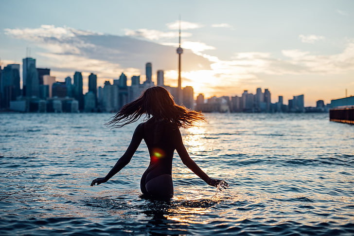 Silhouette of a woman wading into water at sunset with a city skyline and tower in the background, warm golden tones